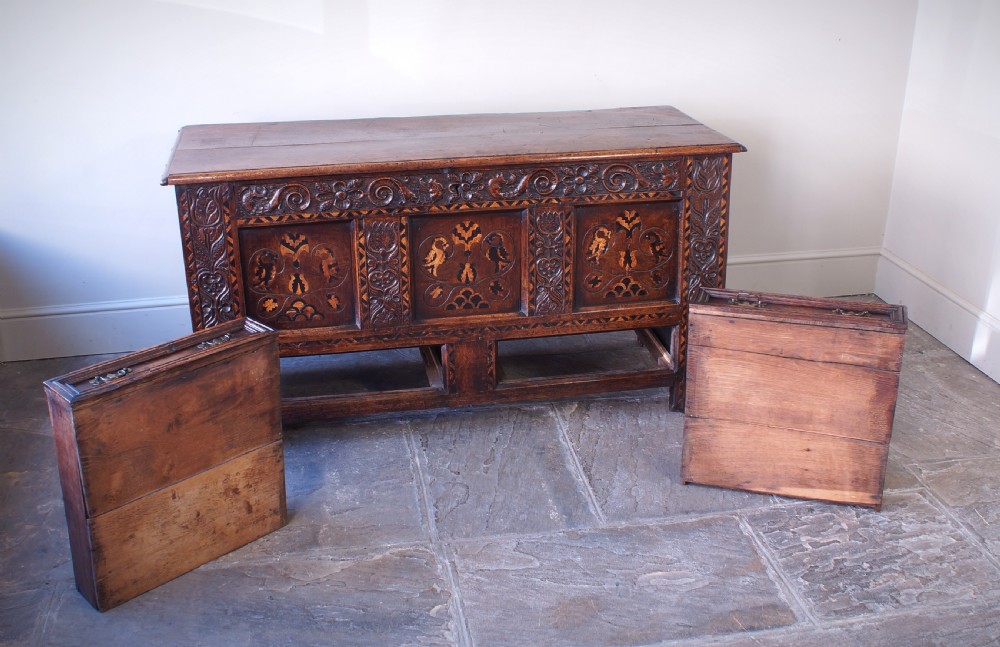 A 17th-century dark oak mule chest featuring carved friezes, three inlaid marquetry front panels with bird motifs, and two lower drawers with brass drop handles. View VI