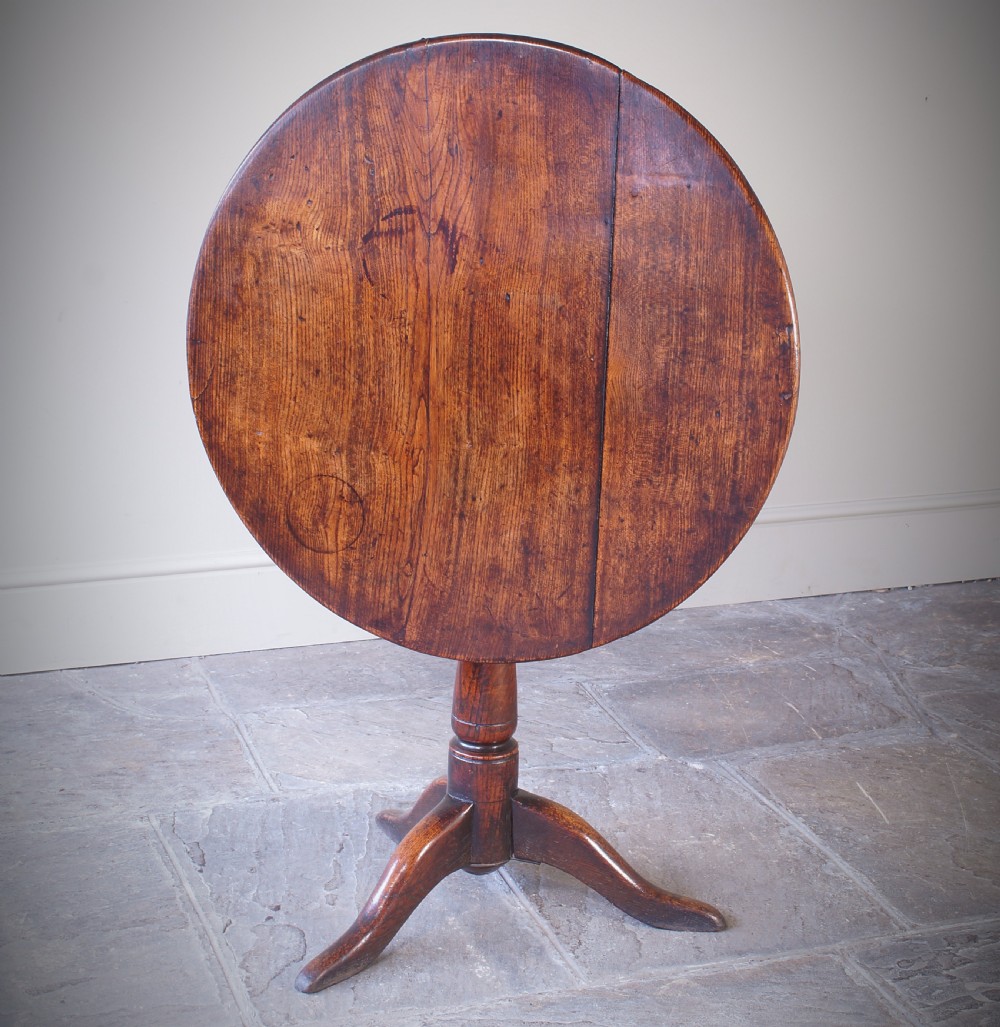 An 18th-century circular elm tilt-top tripod table shown with its top upright on a turned central column and three cabriole legs.