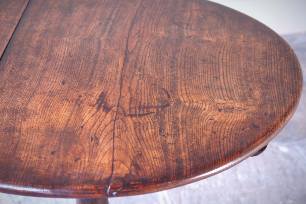 An 18th-century circular elm tilt-top tripod table shown with its top upright on a turned central column and three cabriole legs. View II