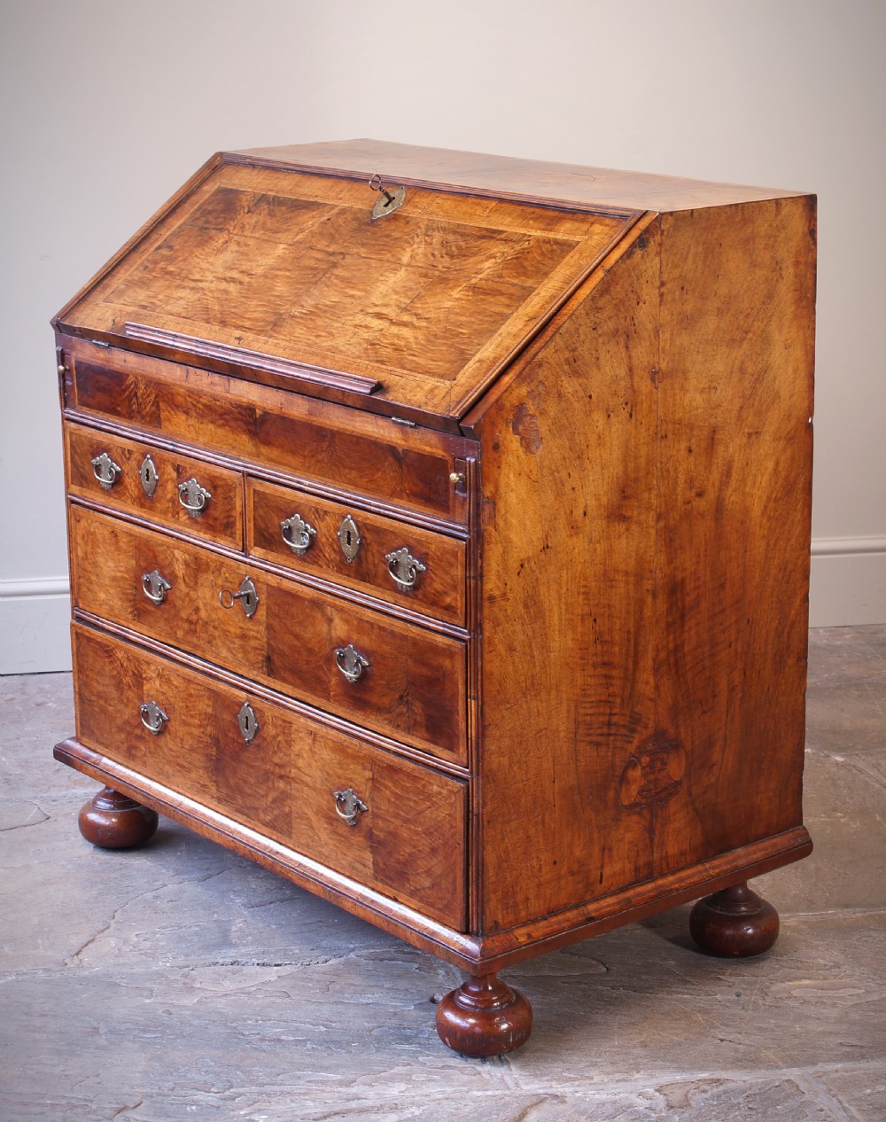An early 18th-century walnut writing bureau featuring a sloping drop-front lid, brass drop handles, and turned bun feet. View VI