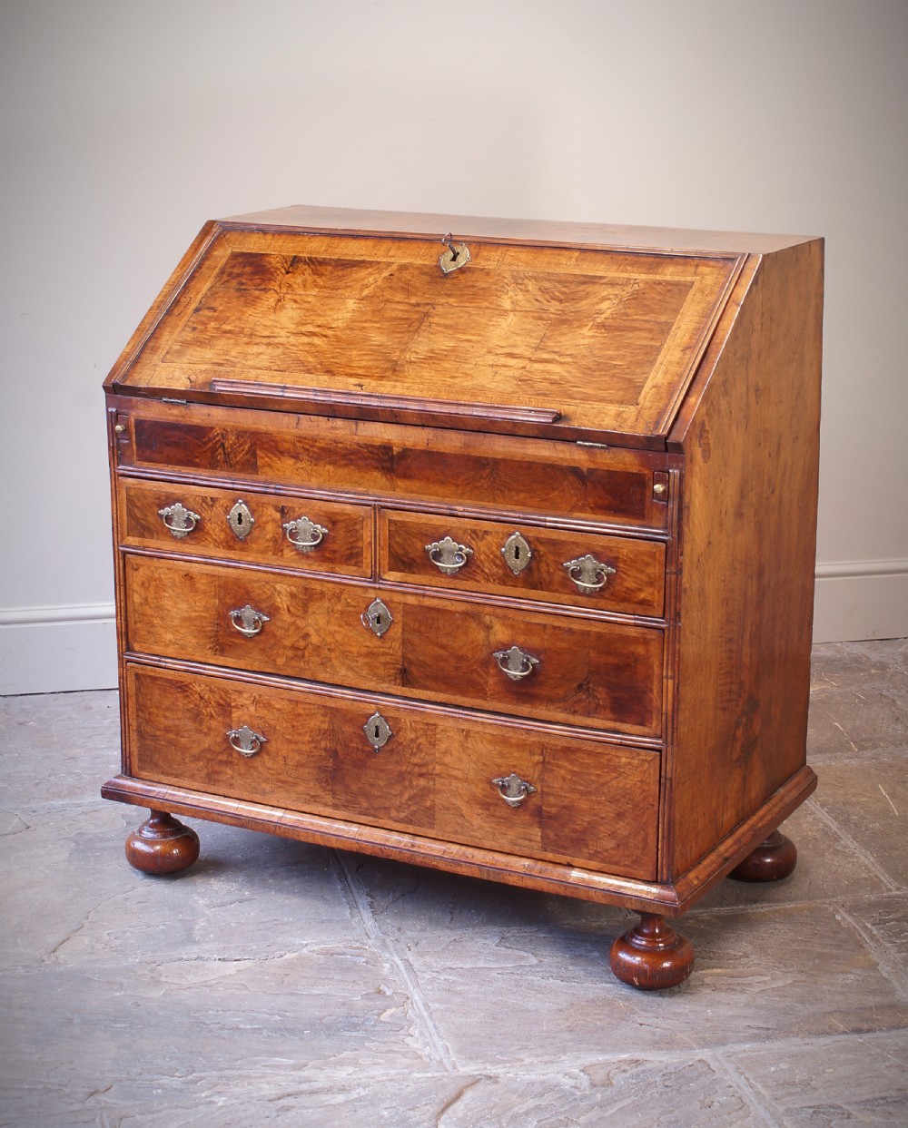 An early 18th-century walnut writing bureau featuring a sloping drop-front lid, brass drop handles, and turned bun feet.