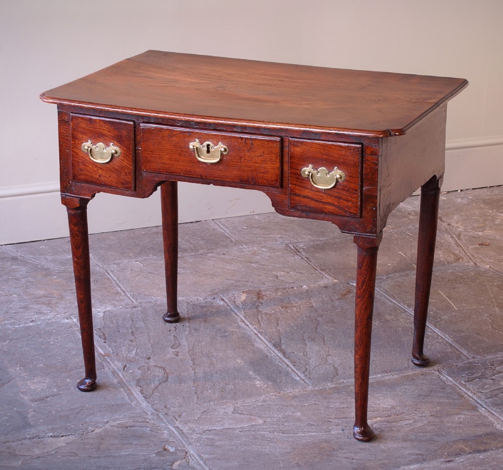 An 18th-century dark brown elm lowboy side table featuring three drawers with brass handles, a shaped apron, and four slender tapered legs ending in pad feet.