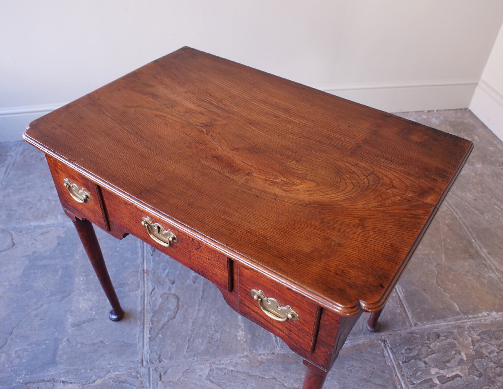 An 18th-century dark brown elm lowboy side table featuring three drawers with brass handles, a shaped apron, and four slender tapered legs ending in pad feet. View II