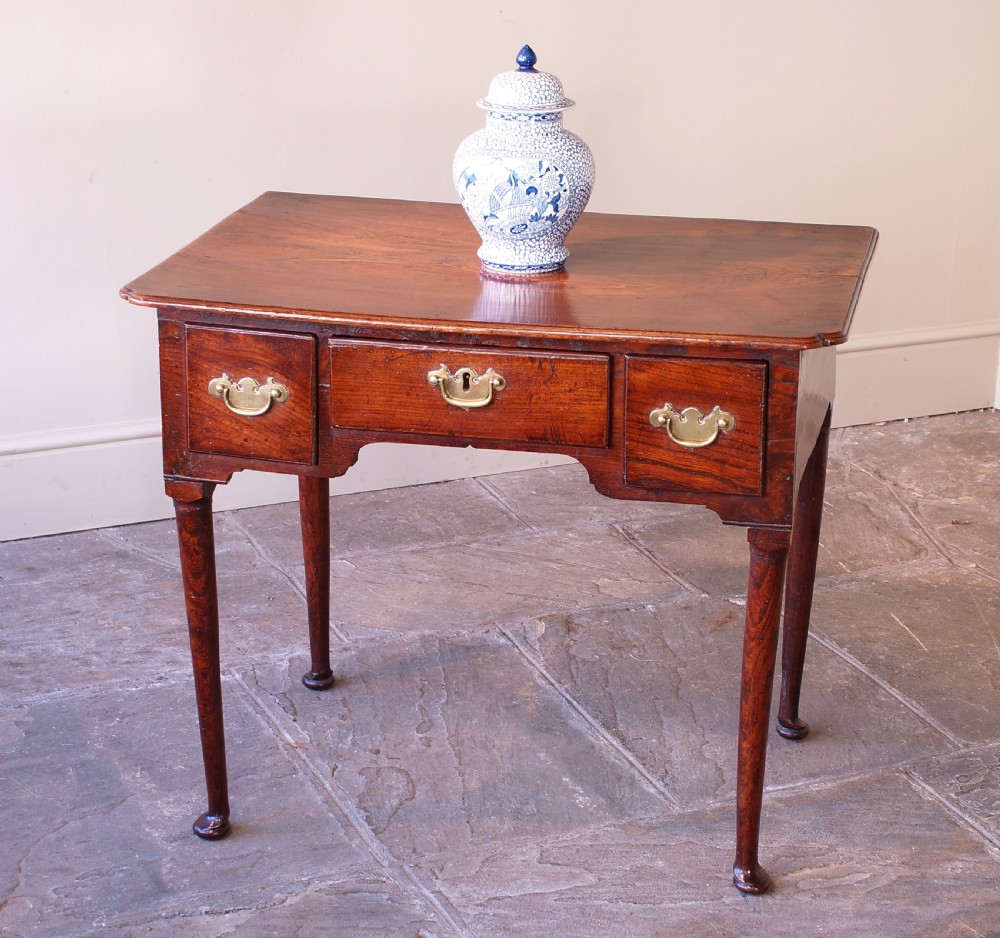 An 18th-century dark brown elm lowboy side table featuring three drawers with brass handles, a shaped apron, and four slender tapered legs ending in pad feet. View VII