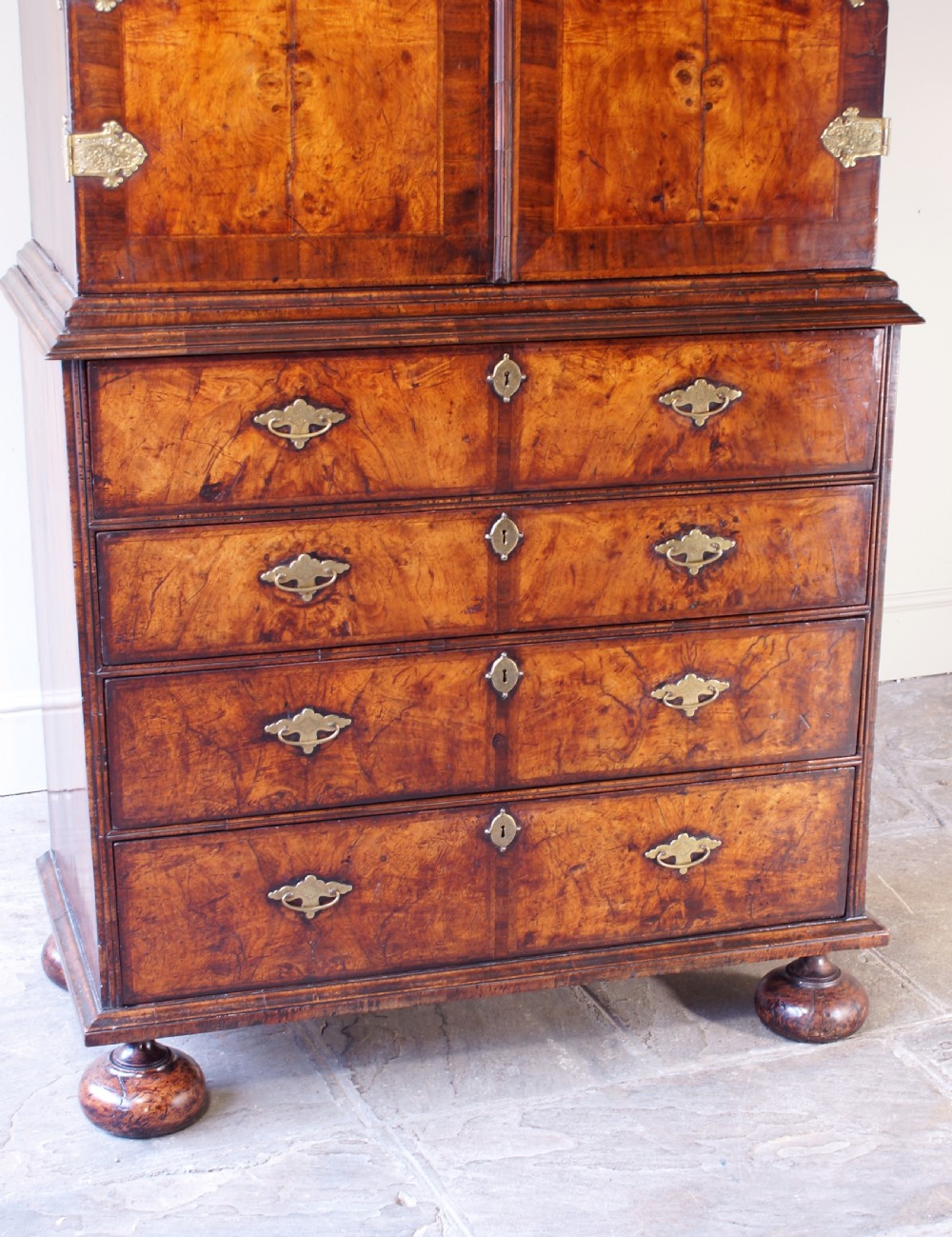 An early eighteenth-century burr walnut cabinet on chest featuring double upper doors with brass strap hinges above four graduated drawers, resting on turned bun feet. View XXV