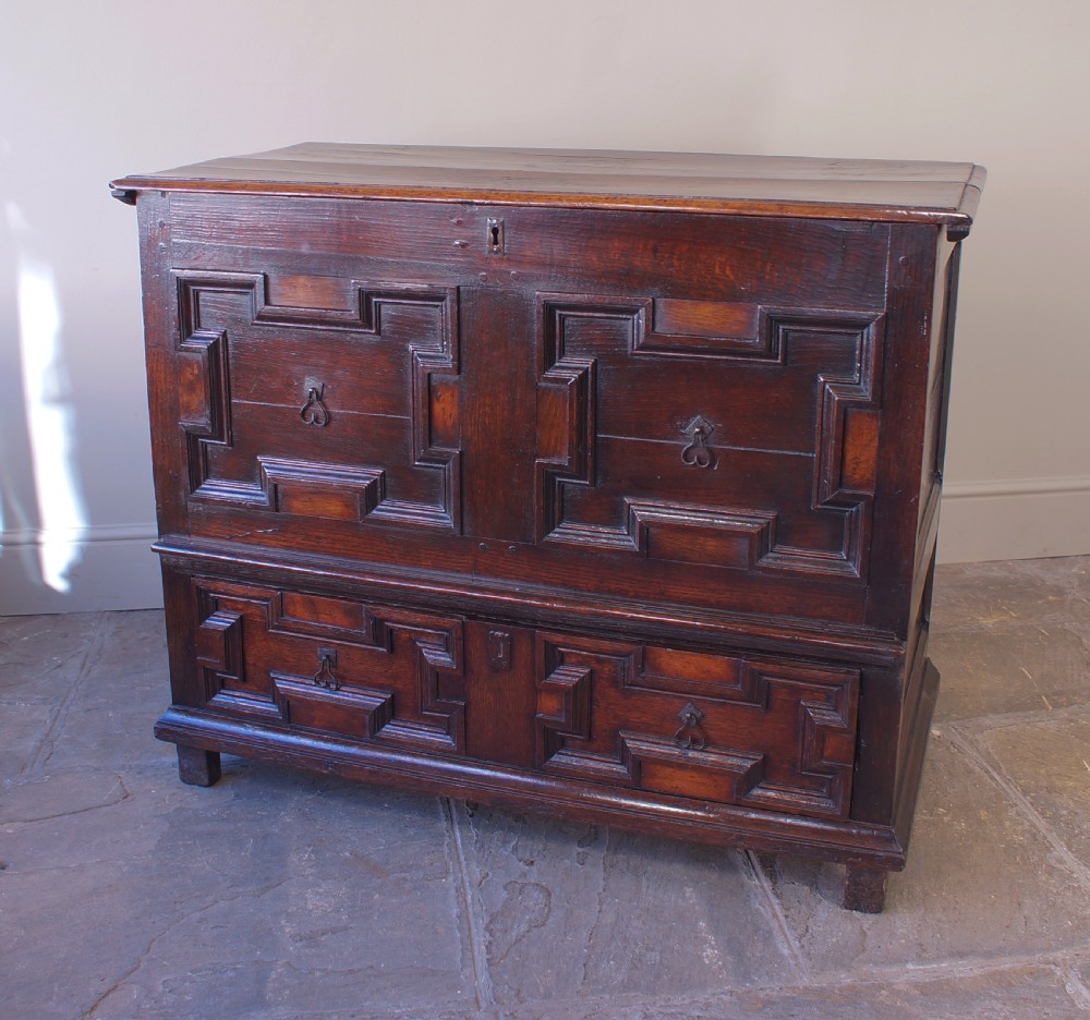 A seventeenth-century dark oak mule chest featuring raised geometric moulded panels on the front lid and two drawers below, set on short block feet.