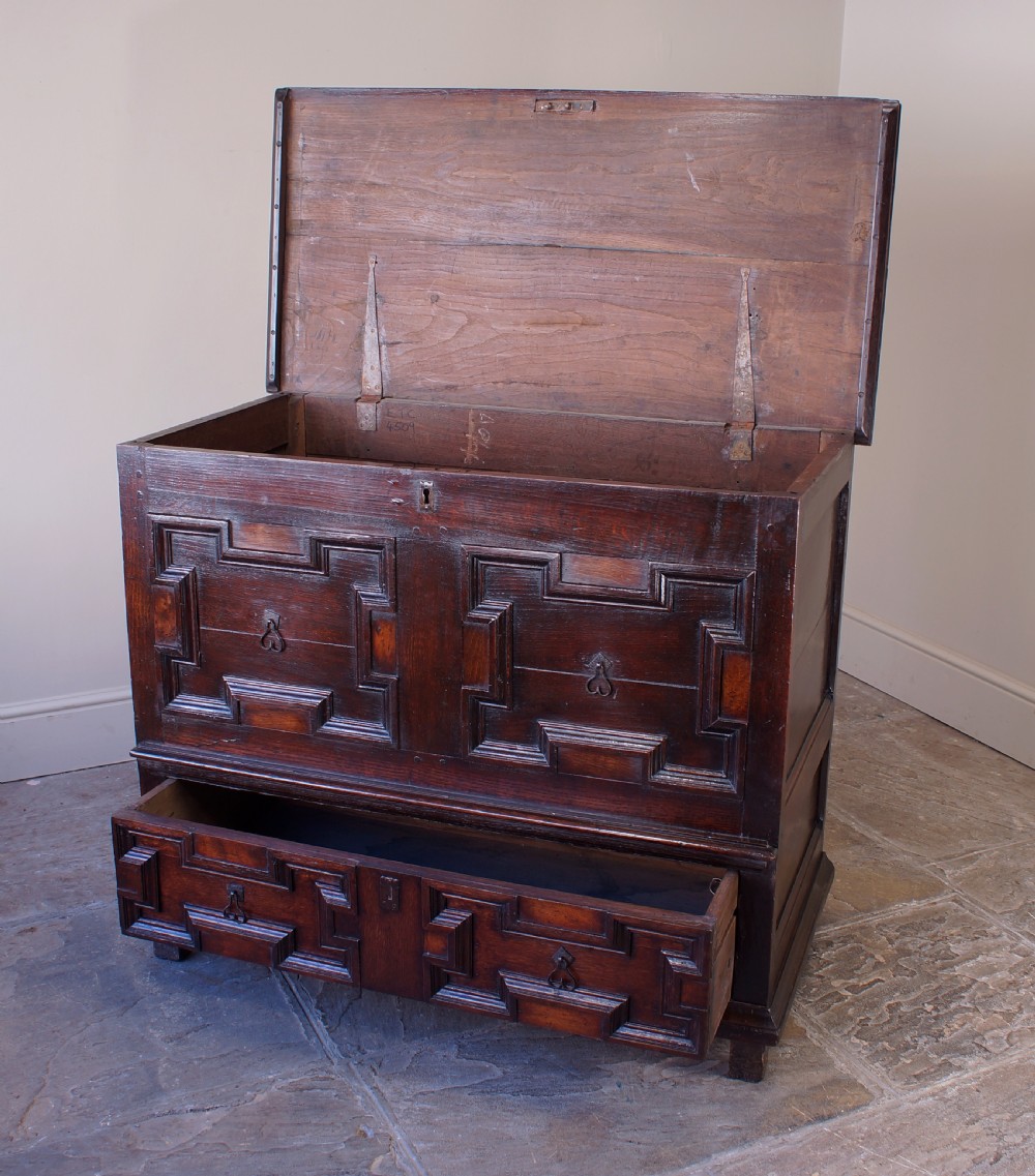 A seventeenth-century dark oak mule chest featuring raised geometric moulded panels on the front lid and two drawers below, set on short block feet. View VI