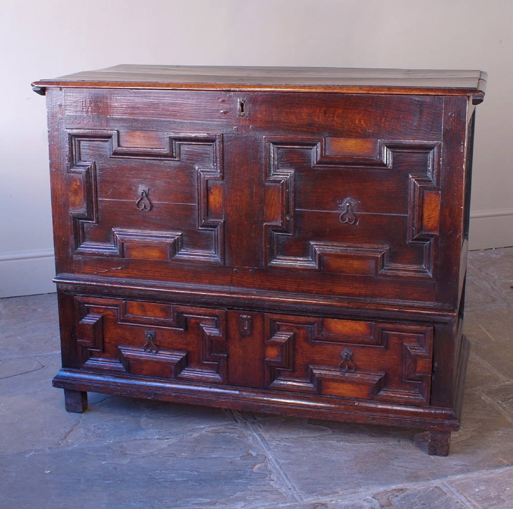 A seventeenth-century dark oak mule chest featuring raised geometric moulded panels on the front lid and two drawers below, set on short block feet. View XI