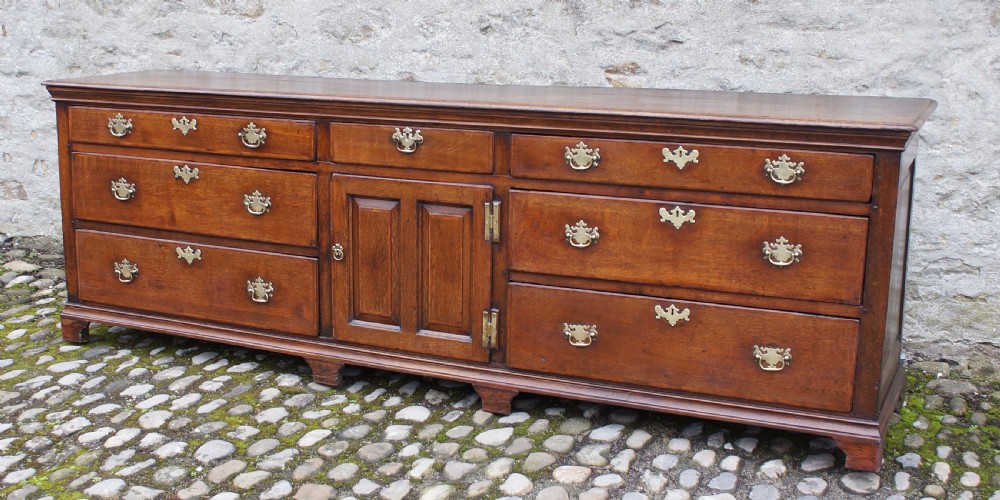 An 18th-century oak dresser base featuring multiple drawers, a central panelled cupboard door, brass handles, and bracket feet. View II