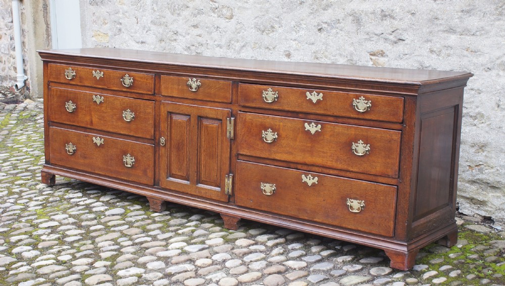 An 18th-century oak dresser base featuring multiple drawers, a central panelled cupboard door, brass handles, and bracket feet. View III