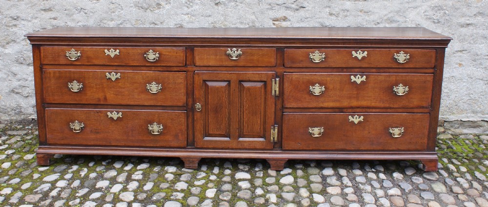 An 18th-century oak dresser base featuring multiple drawers, a central panelled cupboard door, brass handles, and bracket feet. View IV