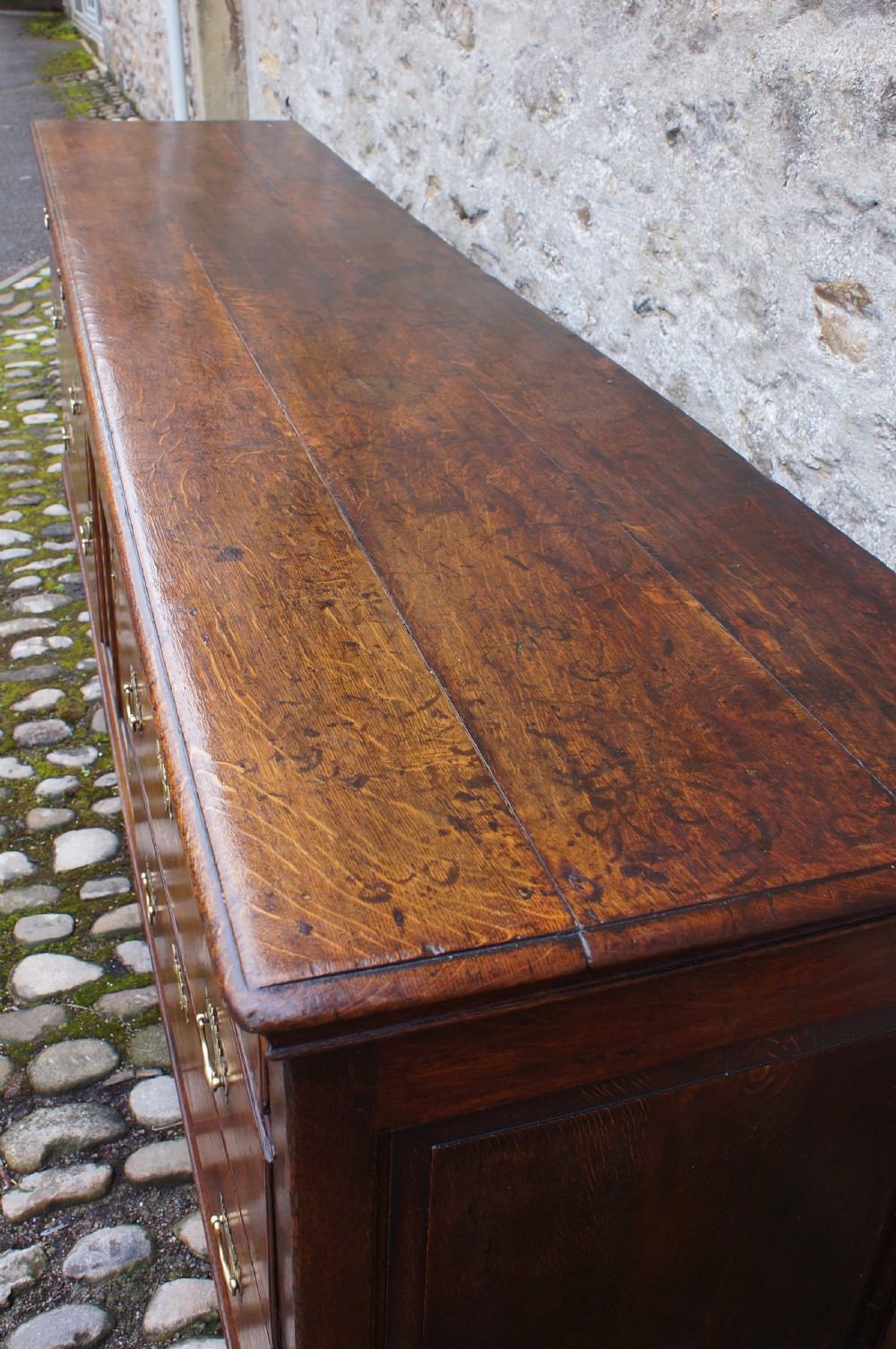 An 18th-century oak dresser base featuring multiple drawers, a central panelled cupboard door, brass handles, and bracket feet. View V