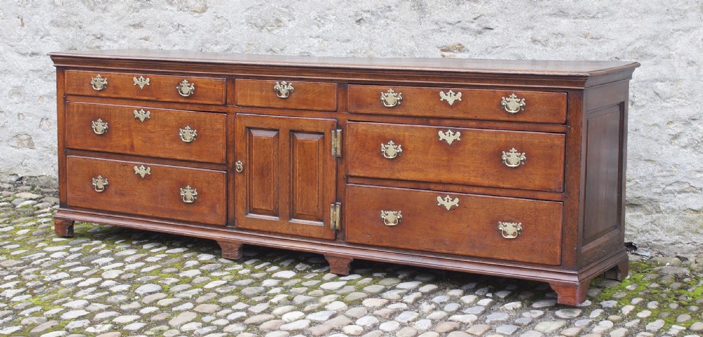 An 18th-century oak dresser base featuring multiple drawers, a central panelled cupboard door, brass handles, and bracket feet. View VI