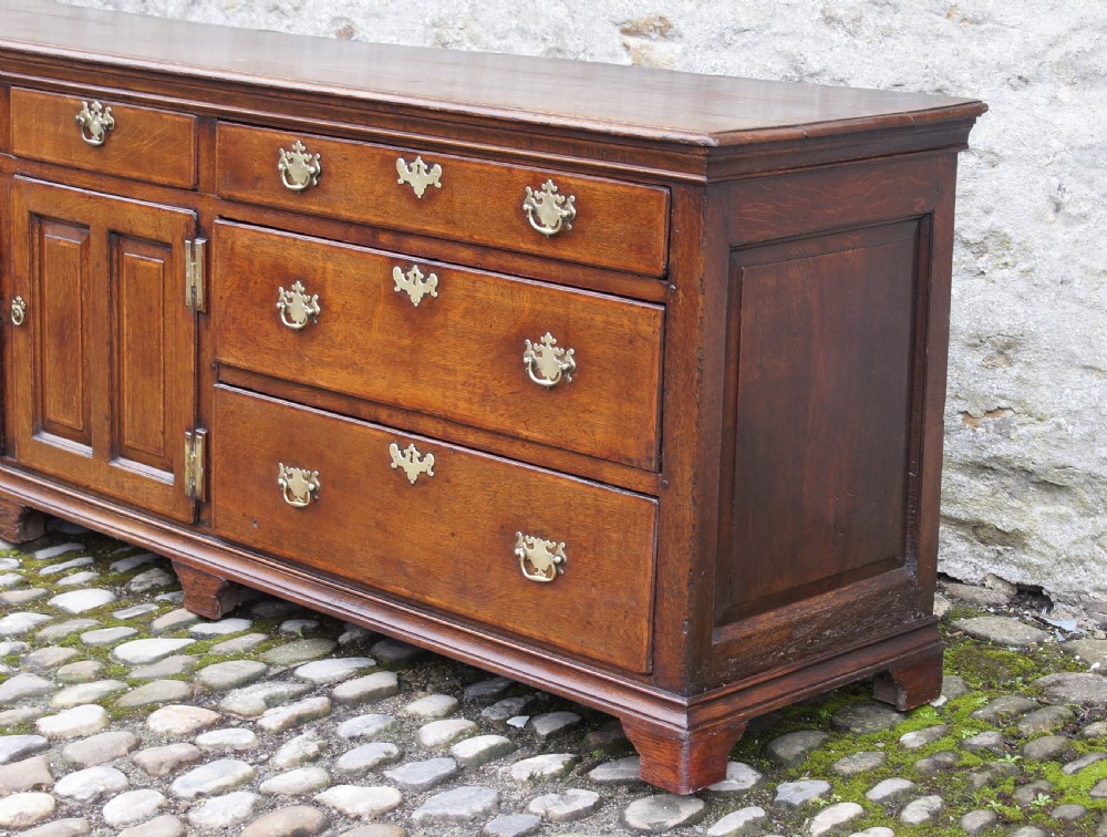 An 18th-century oak dresser base featuring multiple drawers, a central panelled cupboard door, brass handles, and bracket feet. View VII
