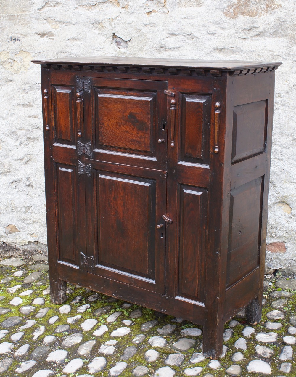 A dark oak 17th century hutch featuring panelled doors, hand-forged iron strap hinges, applied split turnings, and top edge dentil moulding. View III