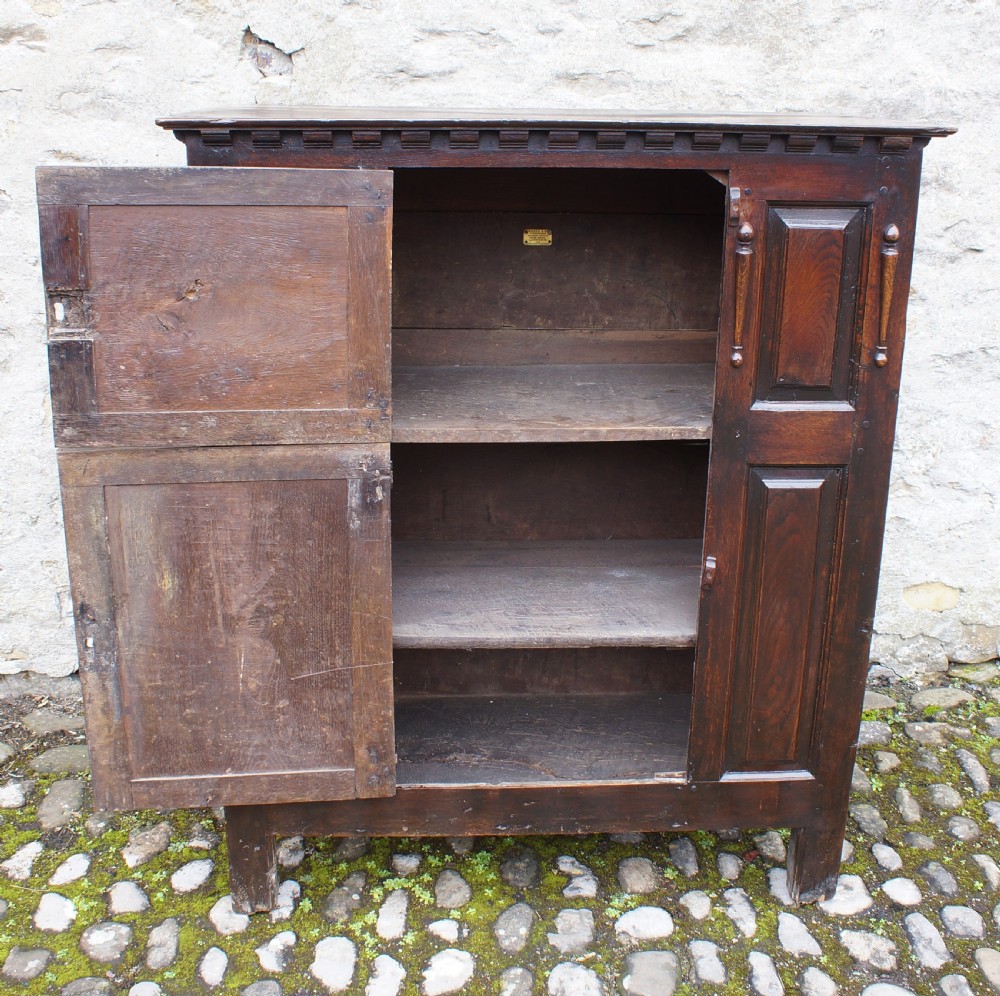 A dark oak 17th century hutch featuring panelled doors, hand-forged iron strap hinges, applied split turnings, and top edge dentil moulding. View V