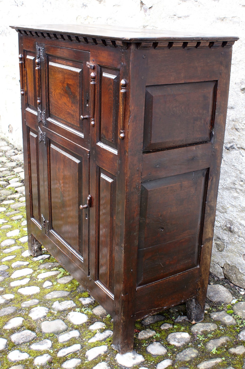 A dark oak 17th century hutch featuring panelled doors, hand-forged iron strap hinges, applied split turnings, and top edge dentil moulding. View VI