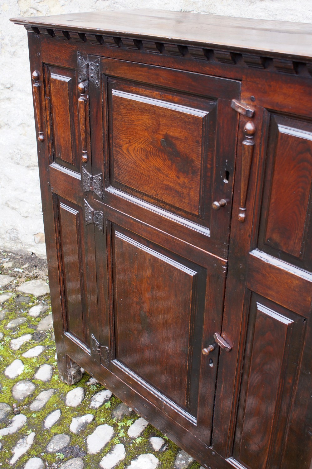 A dark oak 17th century hutch featuring panelled doors, hand-forged iron strap hinges, applied split turnings, and top edge dentil moulding. View VII