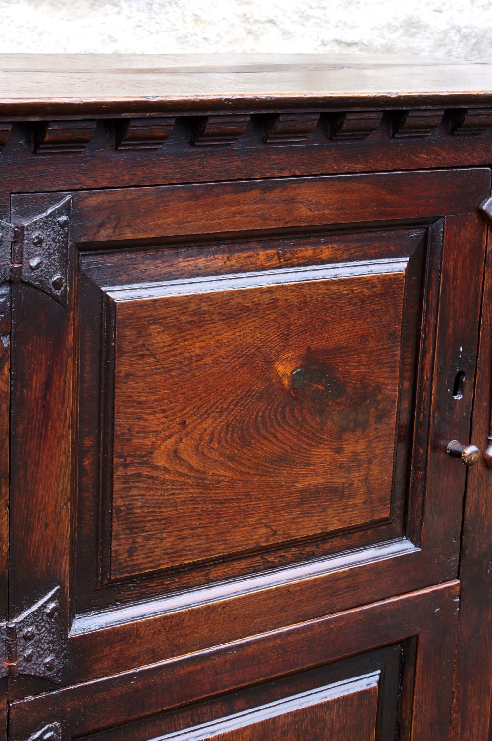 A dark oak 17th century hutch featuring panelled doors, hand-forged iron strap hinges, applied split turnings, and top edge dentil moulding. View XVII