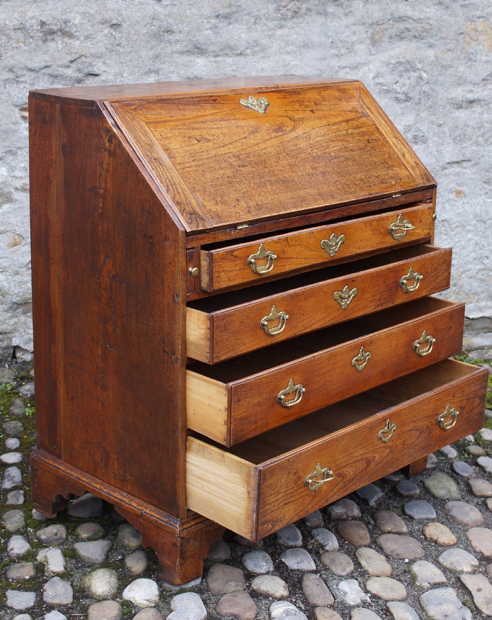An early eighteenth-century elm slant-front bureau featuring four graduated drawers with brass drop handles and bracket feet. View V