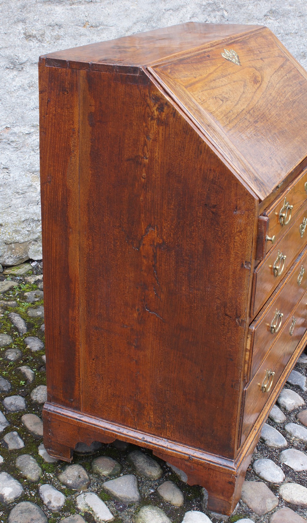 An early eighteenth-century elm slant-front bureau featuring four graduated drawers with brass drop handles and bracket feet. View X