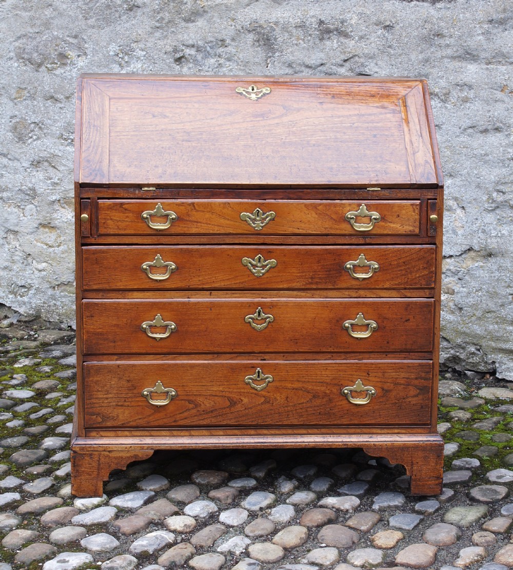 An early eighteenth-century elm slant-front bureau featuring four graduated drawers with brass drop handles and bracket feet. View XV
