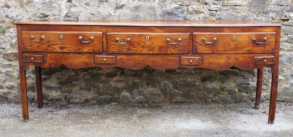 An eighteenth-century warm brown elm dresser base featuring three primary drawers with brass handles above four small drawers set into a scalloped apron. View III