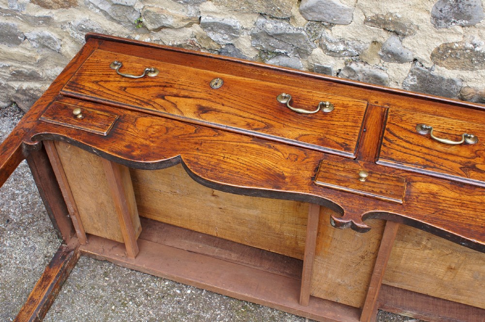 An eighteenth-century warm brown elm dresser base featuring three primary drawers with brass handles above four small drawers set into a scalloped apron. View XVIII