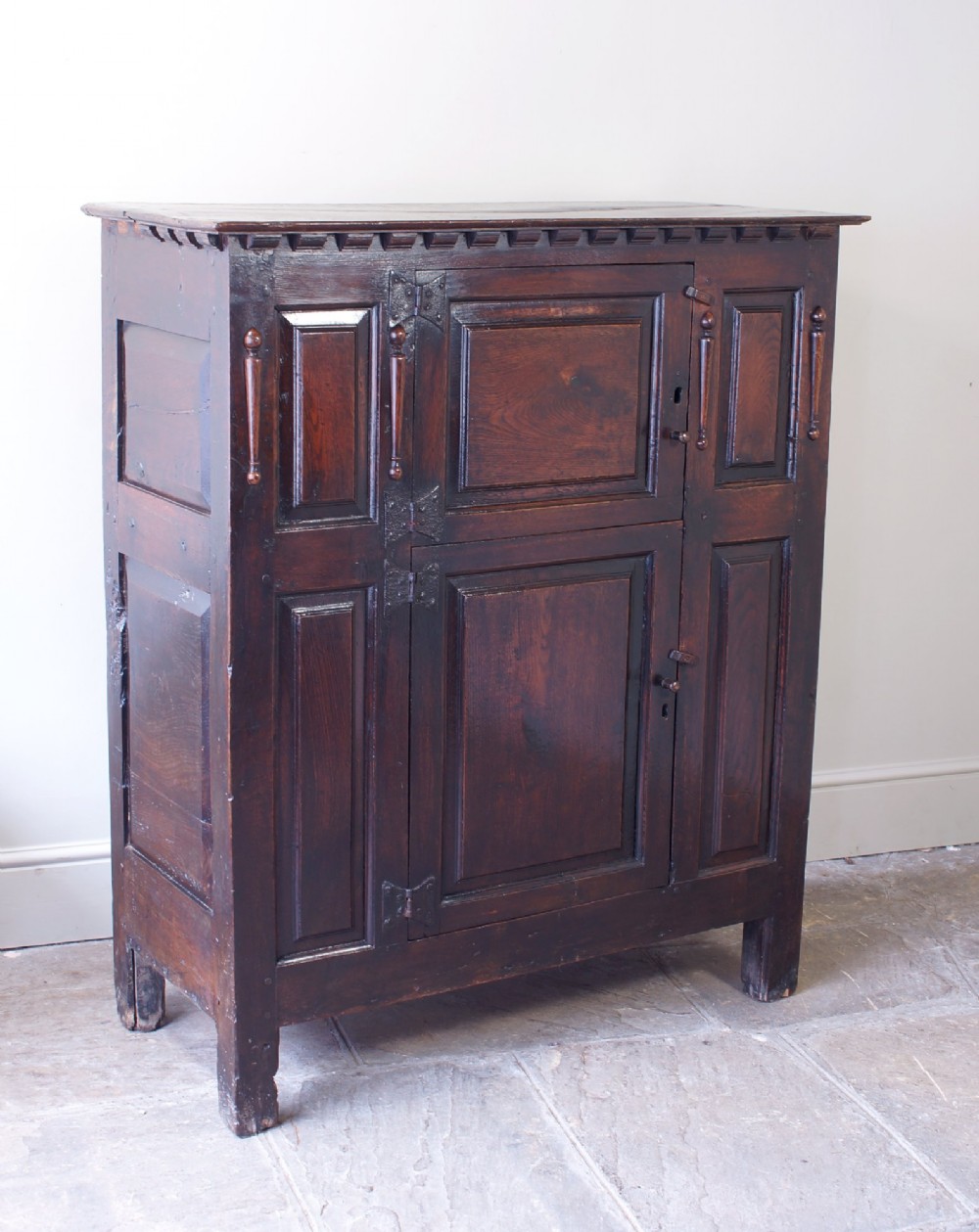 A dark oak 17th century hutch featuring panelled doors, hand-forged iron strap hinges, applied split turnings, and top edge dentil moulding. View XIV
