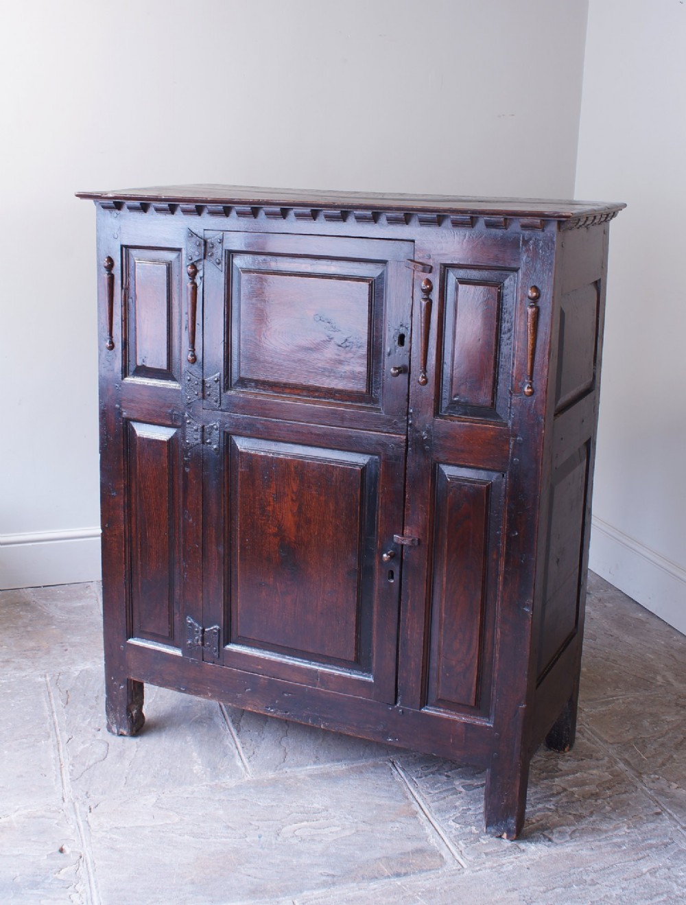 A dark oak 17th century hutch featuring panelled doors, hand-forged iron strap hinges, applied split turnings, and top edge dentil moulding. View XV