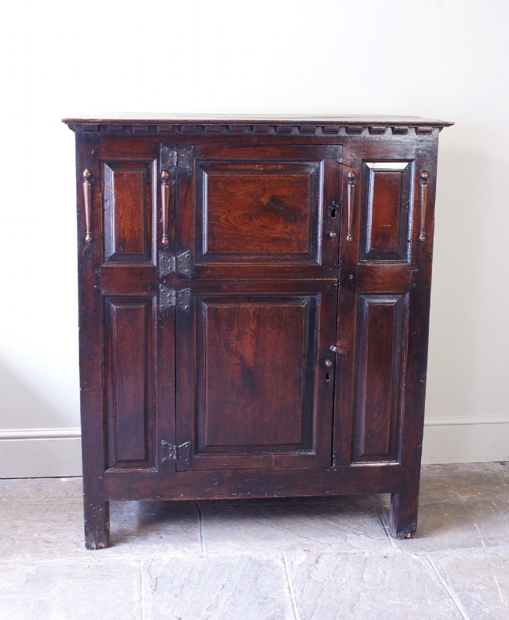 A dark oak 17th century hutch featuring panelled doors, hand-forged iron strap hinges, applied split turnings, and top edge dentil moulding.
