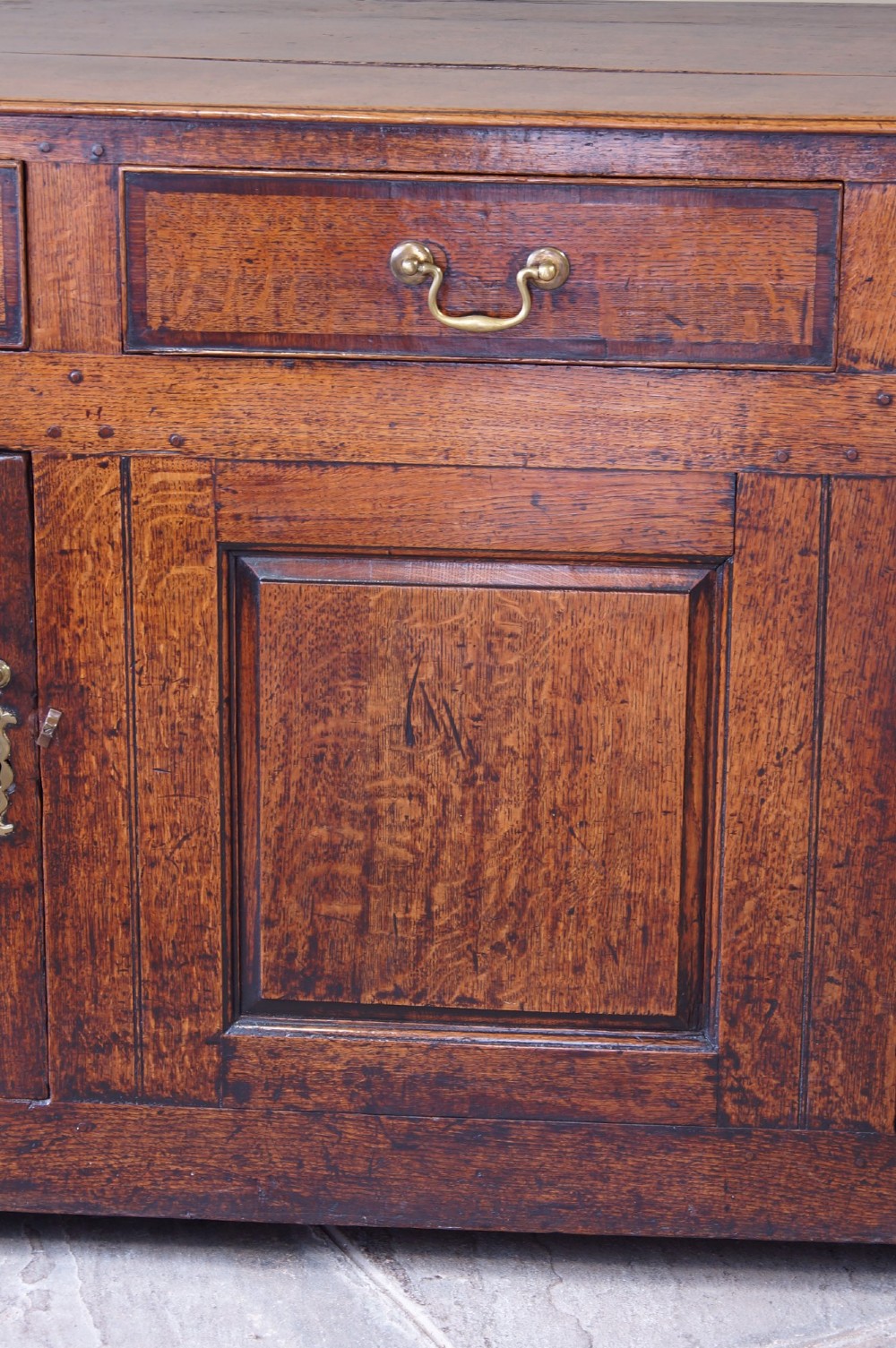 A Georgian oak dresser base featuring three top drawers with brass handles above three panelled cupboard doors. View VI