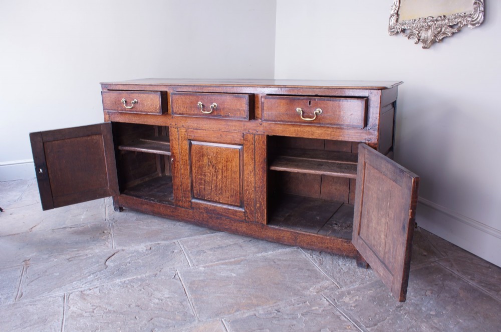 A Georgian oak dresser base featuring three top drawers with brass handles above three panelled cupboard doors. View XI