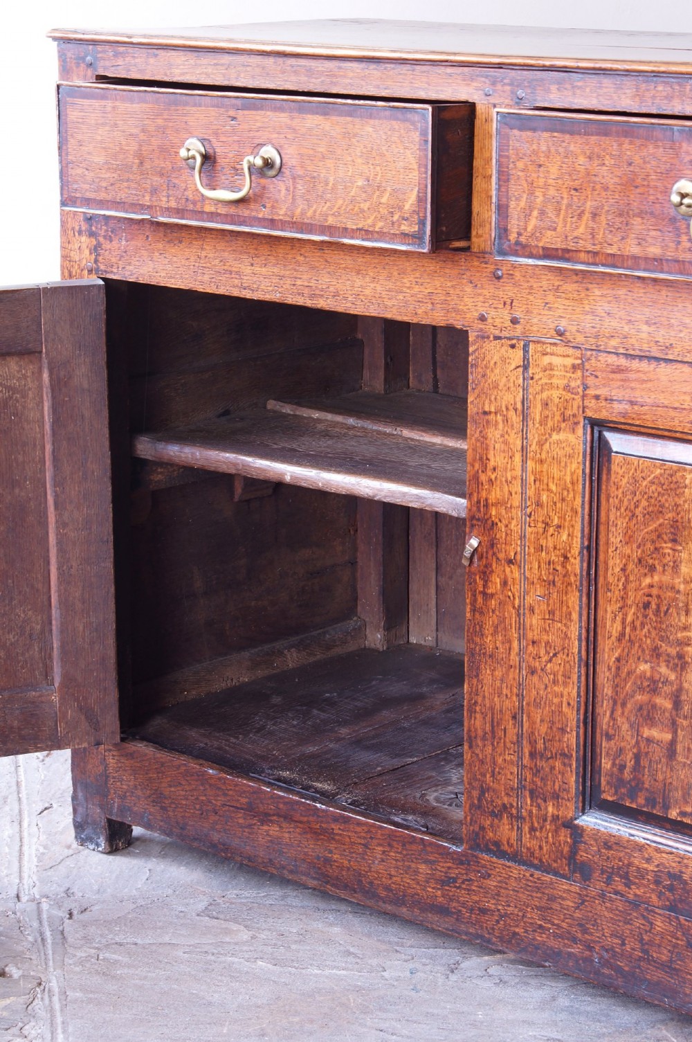 A Georgian oak dresser base featuring three top drawers with brass handles above three panelled cupboard doors. View XIV