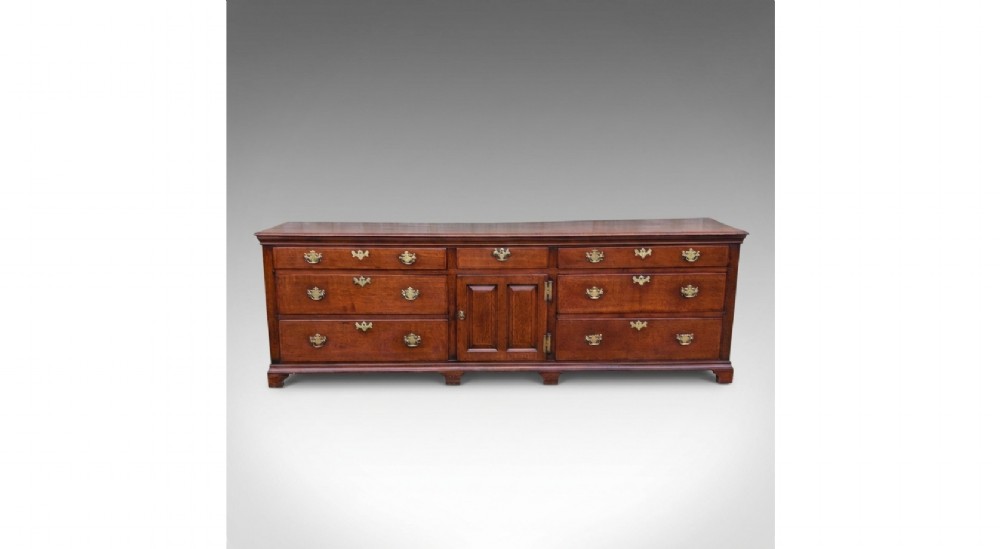 An 18th-century oak dresser base featuring multiple drawers, a central panelled cupboard door, brass handles, and bracket feet.