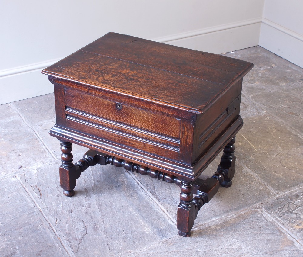 A 17th-century dark oak box stool with a flat hinged top, moulded panel front, and turned legs joined by a bobbin stretcher. View II
