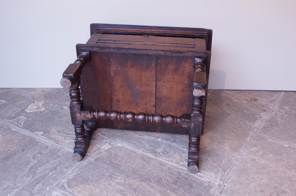 A 17th-century dark oak box stool with a flat hinged top, moulded panel front, and turned legs joined by a bobbin stretcher. View III
