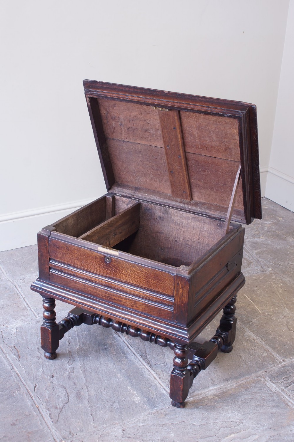 A 17th-century dark oak box stool with a flat hinged top, moulded panel front, and turned legs joined by a bobbin stretcher. View IV
