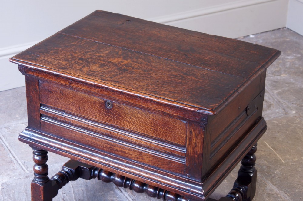 A 17th-century dark oak box stool with a flat hinged top, moulded panel front, and turned legs joined by a bobbin stretcher. View V