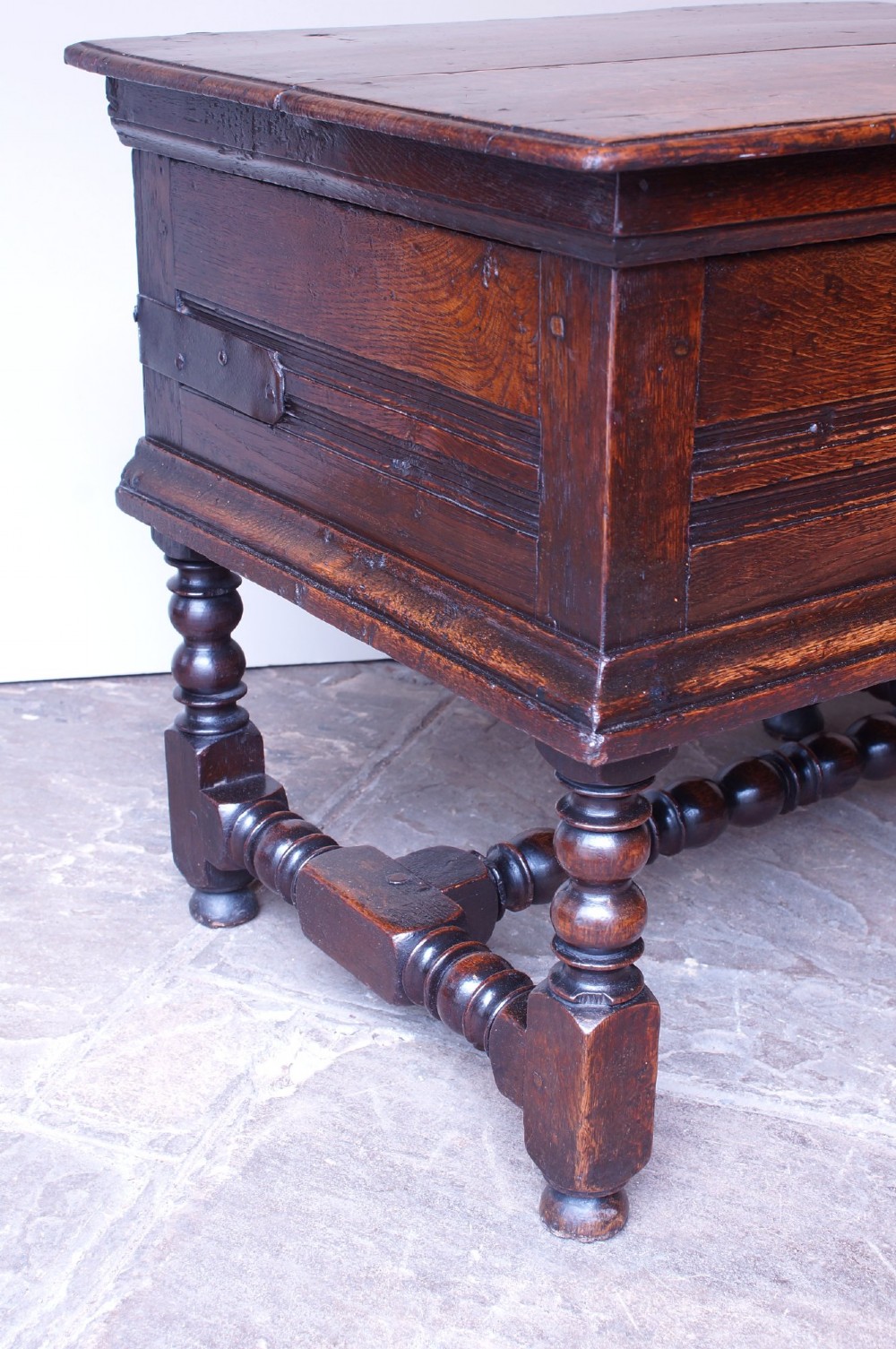 A 17th-century dark oak box stool with a flat hinged top, moulded panel front, and turned legs joined by a bobbin stretcher. View VII