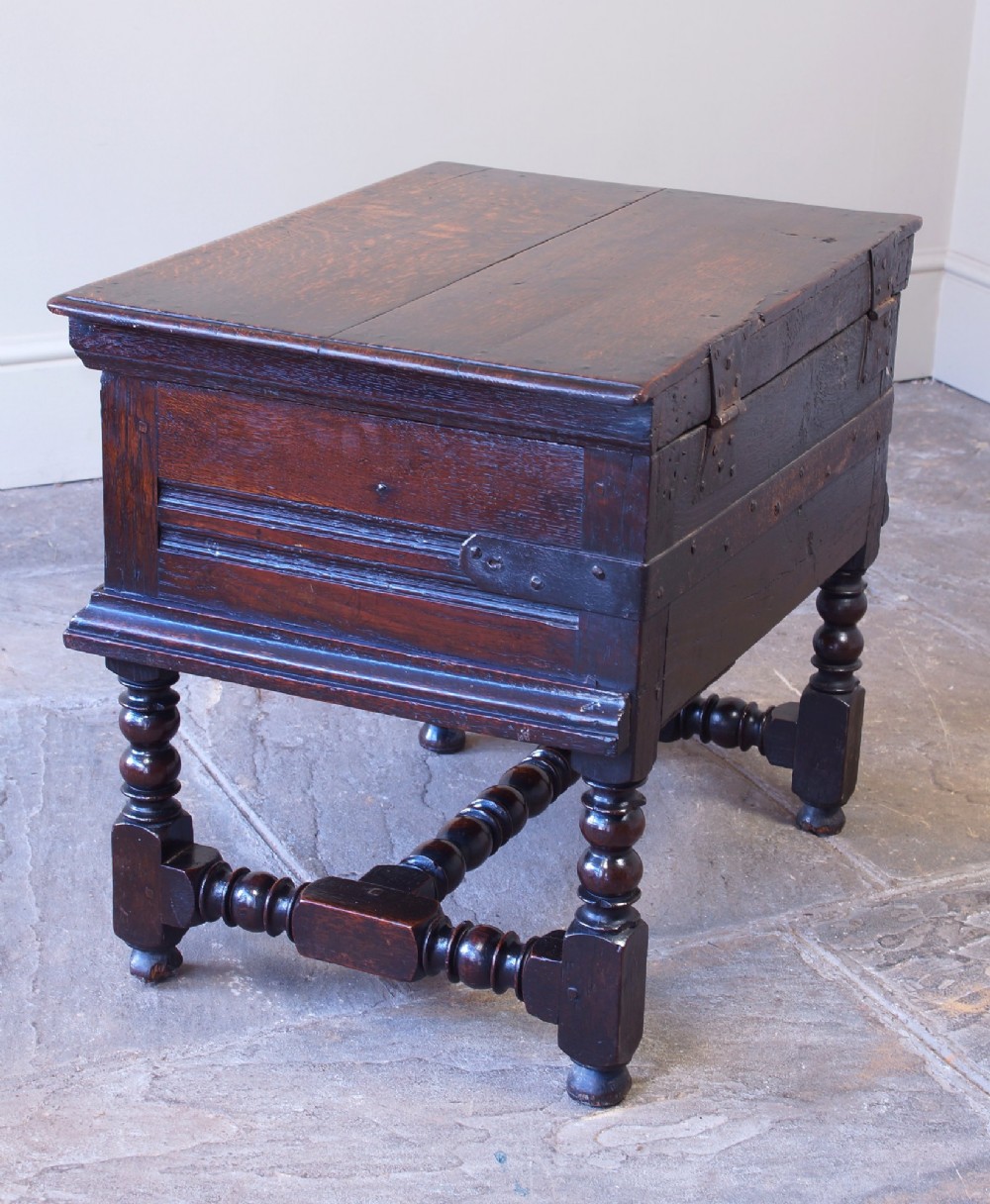 A 17th-century dark oak box stool with a flat hinged top, moulded panel front, and turned legs joined by a bobbin stretcher. View X