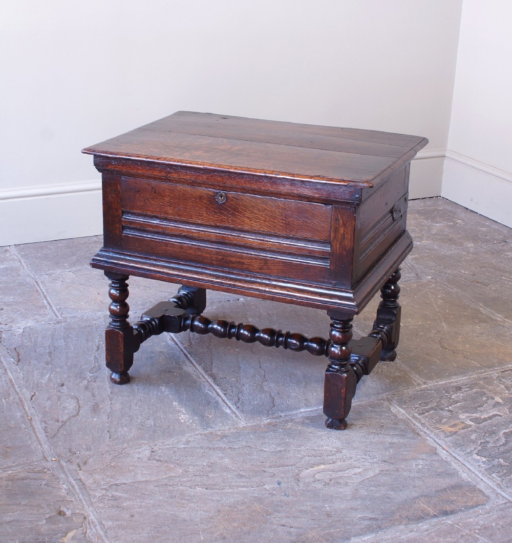 A 17th-century dark oak box stool with a flat hinged top, moulded panel front, and turned legs joined by a bobbin stretcher.