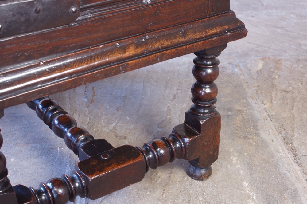 A 17th-century dark oak box stool with a flat hinged top, moulded panel front, and turned legs joined by a bobbin stretcher. View XIV
