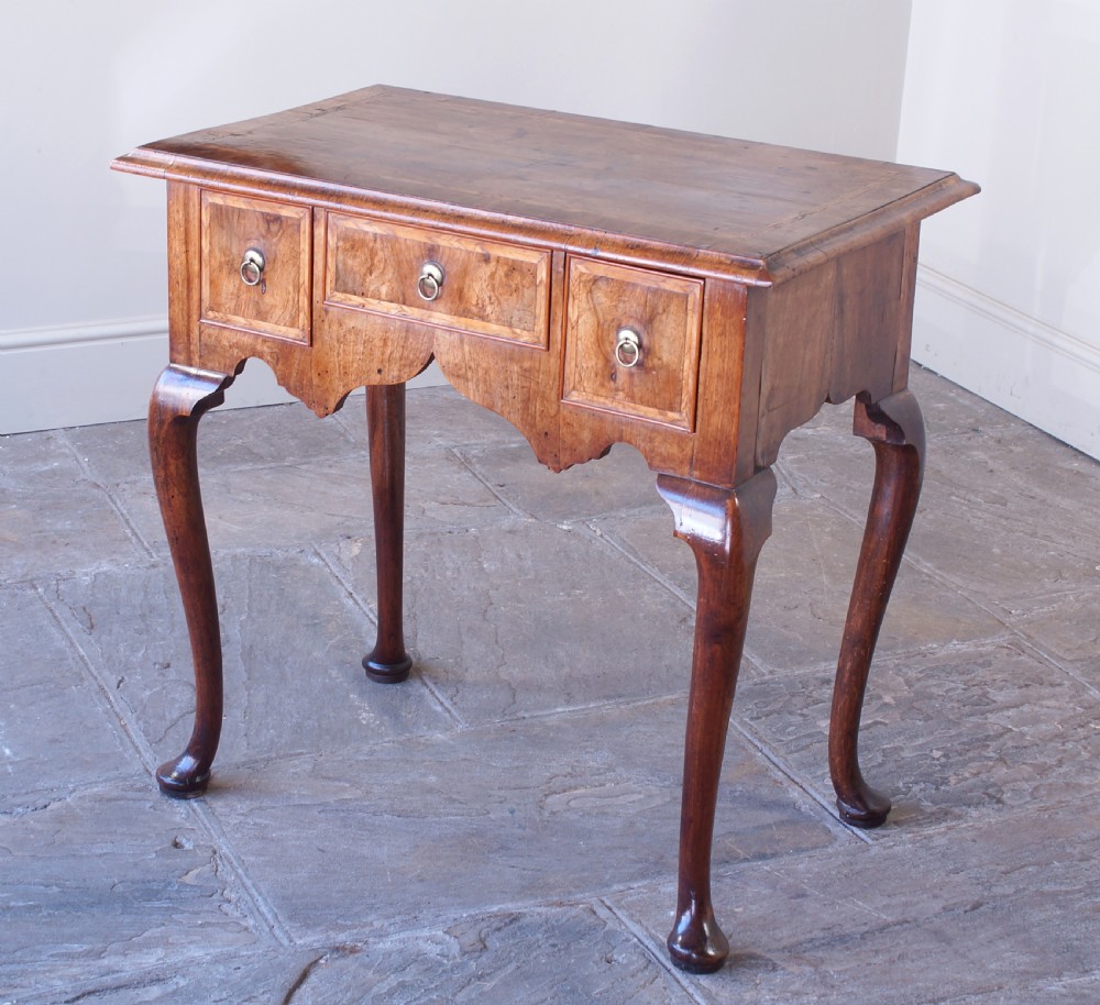 An early 18th-century walnut lowboy table with three drawers, brass ring pull handles, a scalloped apron, and cabriole legs ending in pad feet.