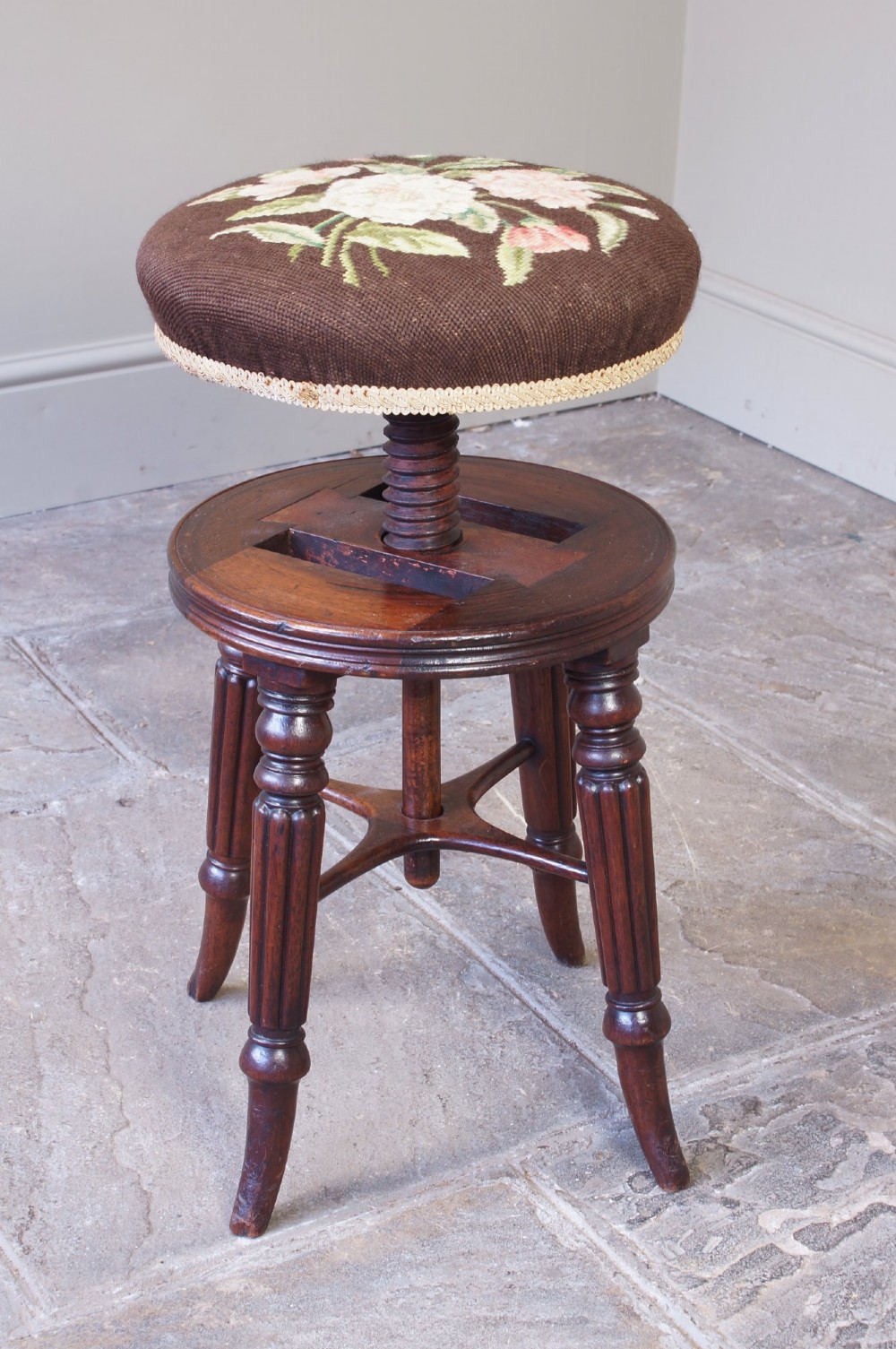 A Regency mahogany revolving piano stool featuring a circular floral needlepoint cushion, central wooden screw mechanism, and fluted turned legs. View V