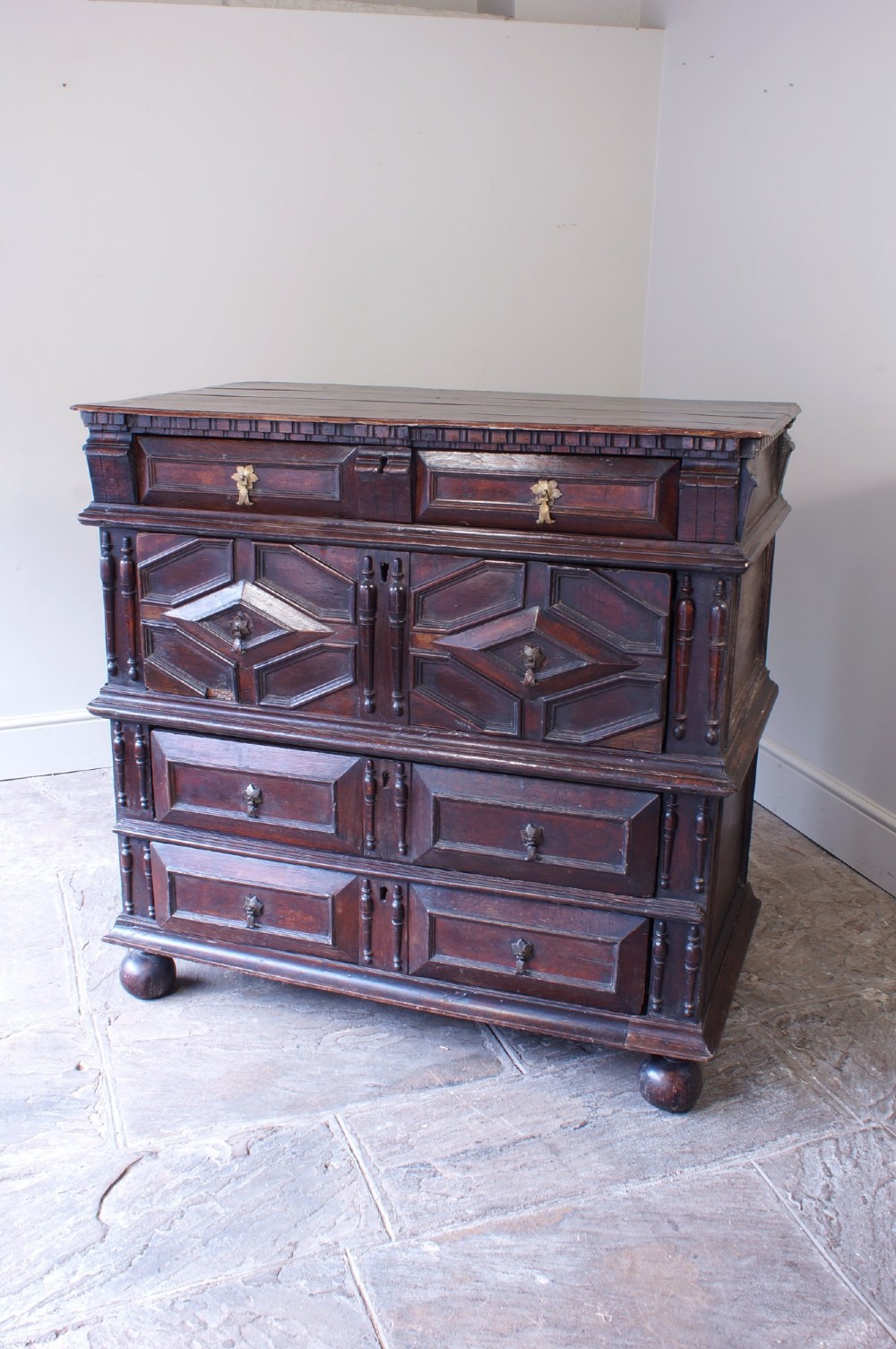 A 17th-century Charles II dark oak chest of drawers featuring geometric mitred panels, turned split spindles, brass drop handles, and bun feet.