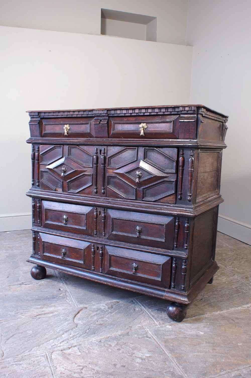 A 17th-century Charles II dark oak chest of drawers featuring geometric mitred panels, turned split spindles, brass drop handles, and bun feet. View VI