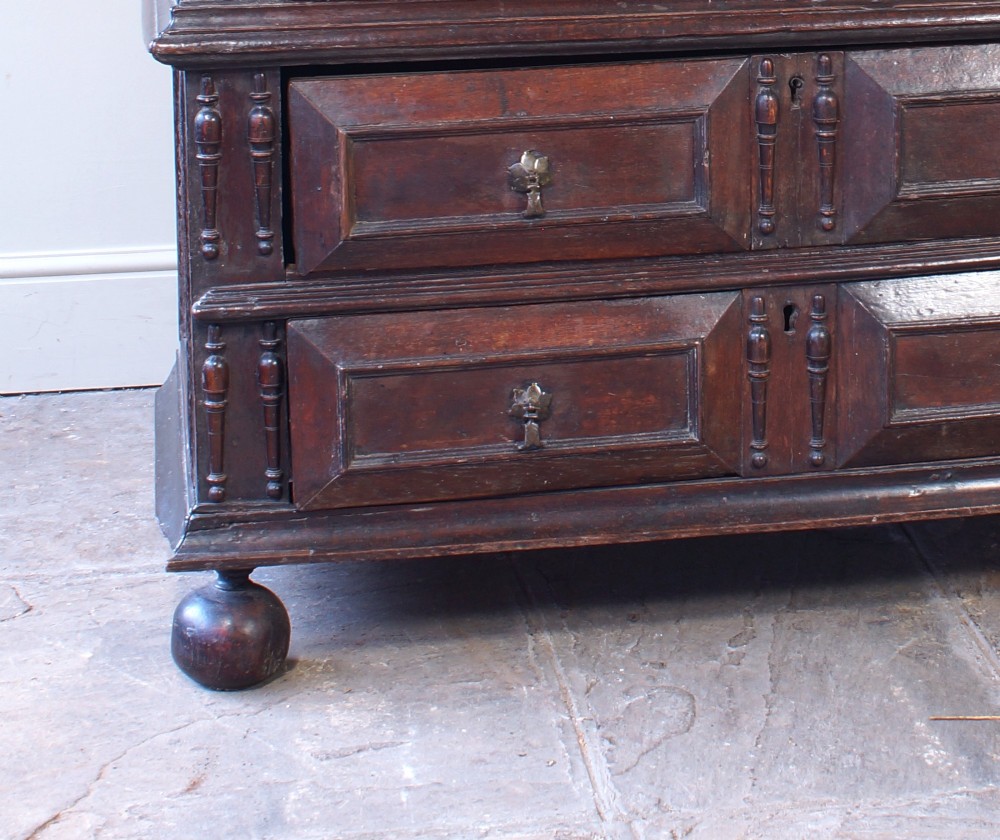 A 17th-century Charles II dark oak chest of drawers featuring geometric mitred panels, turned split spindles, brass drop handles, and bun feet. View VII
