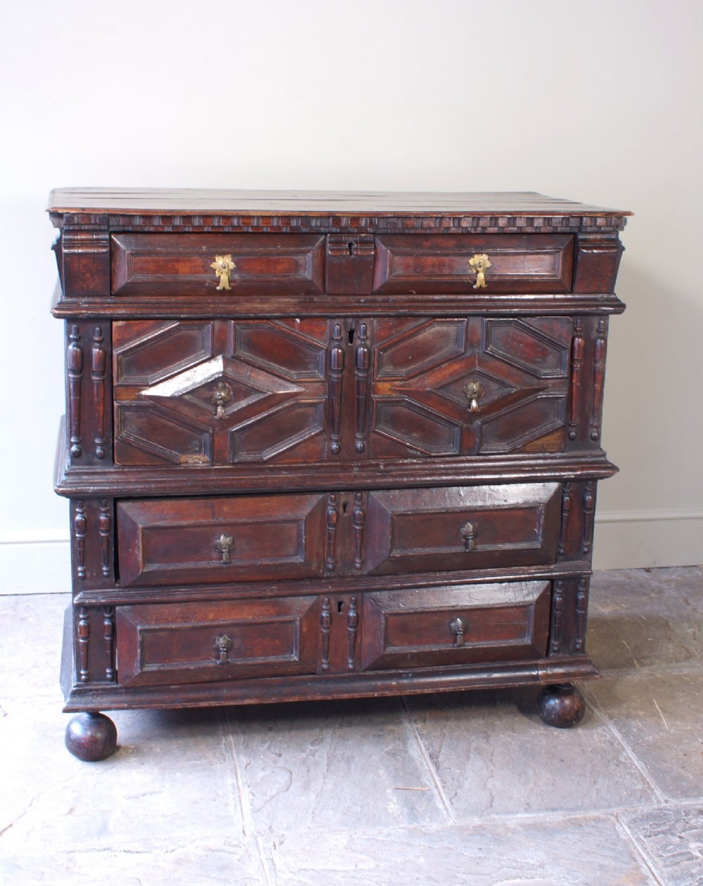 A 17th-century Charles II dark oak chest of drawers featuring geometric mitred panels, turned split spindles, brass drop handles, and bun feet. View X
