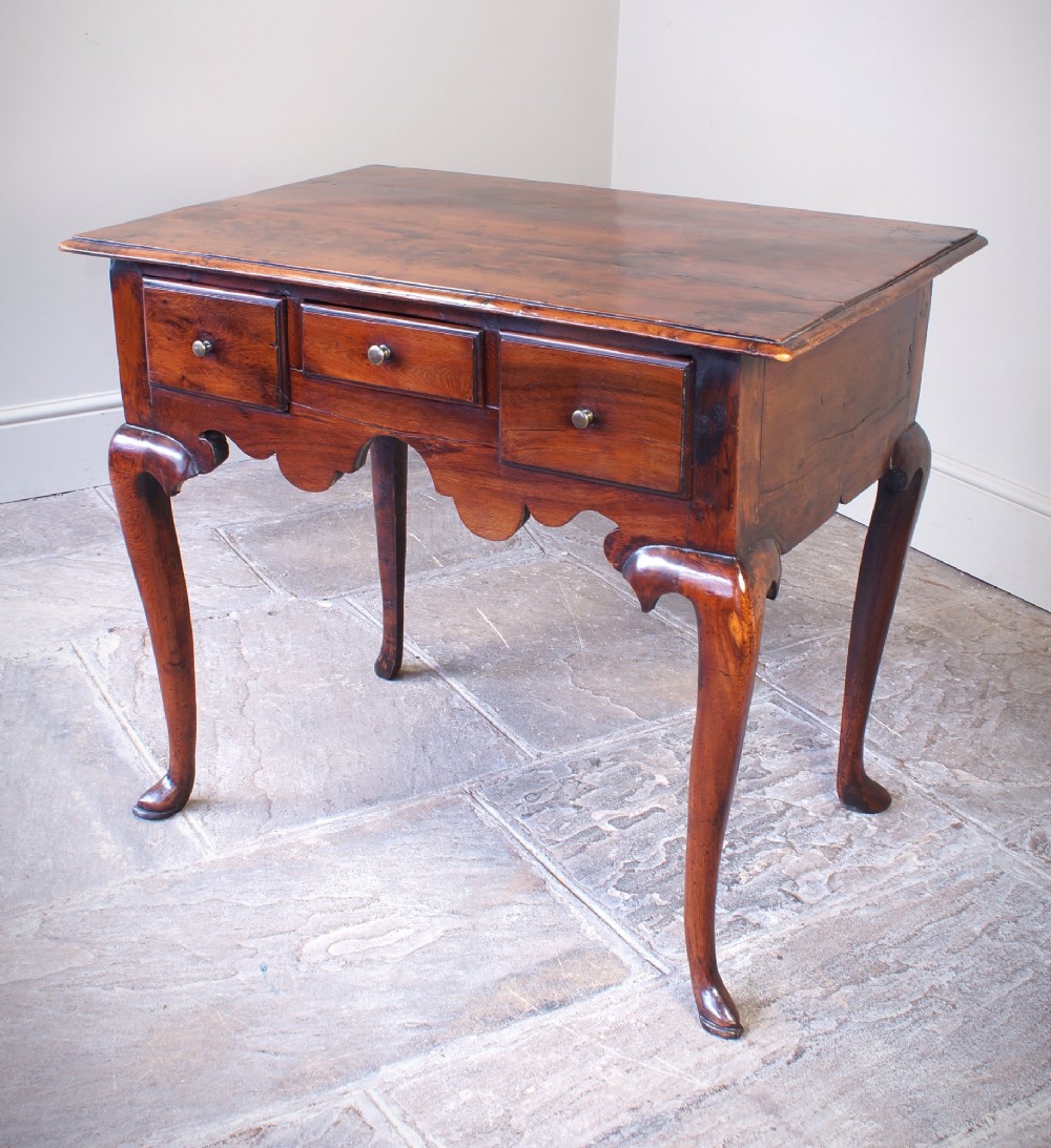 An 18th-century yew wood lowboy table featuring three drawers, a shaped scalloped apron, metal knob handles, and cabriole legs.